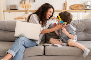 Happy mom and son laugh sit together on sofa: mother work on laptop and kid in headphones listen to...