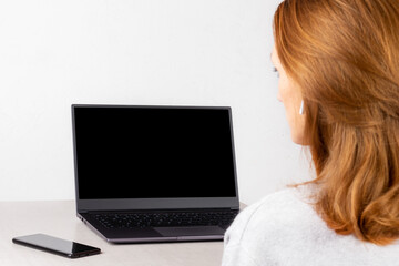 Naklejka premium red-haired young woman sitting in front of a laptop with a black mockup on the screen, concept of distance learning, live broadcast, online communication