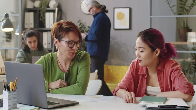 Senior female mentor discussing project on laptop screen with young Asian trainee while helping her during probation period in office