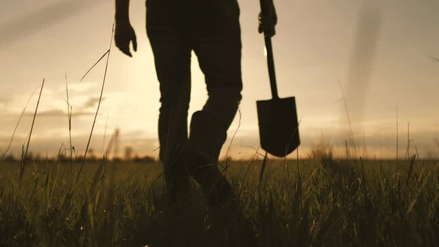 Farming. Young farmer man holding shovel on growing field at sunset. Worker man walks through the black soil with a shovel in his hands. Agriculture harvesting food farmland ground concept.