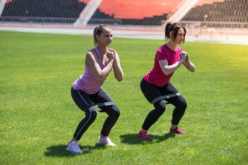 Two women squat on the grass in the stadium. Gymnastics, weight loss, sport