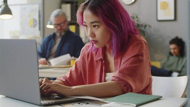Tilt up portrait shot of young Asian female trainee with pink hair using laptop and posing for camera at office desk