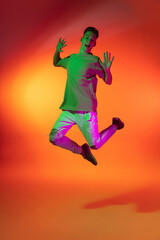 Fototapeta na wymiar Portrait of Caucasian young man jumping, dancing isolated on orange studio background in neon light. Concept of human emotions, facial expression.