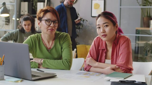 Portrait of senior Caucasian female mentor and young Asian trainee sitting together at office desk and posing for camera