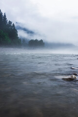 Dark and moody river and rocks landscape with fog in the trees and water. Moody nature vibes with mist and dark cold weather.