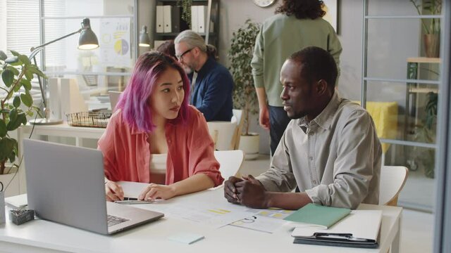 Afro-American male mentor and young Asian female trainee having discussion and using laptop while working together in office