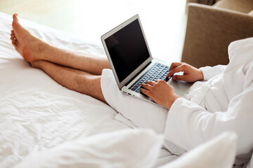 Naklejka premium a man in a robe lies on a bed with a computer on his feet. Freelance, work from home. No face. A man in a hotel, working in a bedroom at a computer.