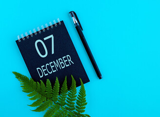 December 7th. Day 7 of month, Calendar date. Black notepad sheet, pen, fern twig, on a blue background. Winter month, day of the year concept