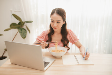 Obraz na płótnie Canvas Girl eating instant noodle in hurry time while running her business at home on notebook computer