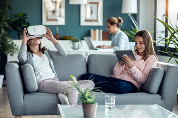 Little girl using virtual reality headset while her mother and sister working in living room at home.