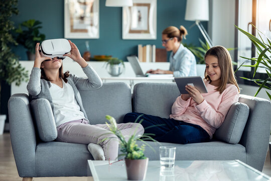 Little girl using virtual reality headset while her mother and sister working in living room at home.