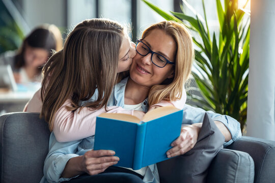Mature beautiful woman reading a book while her daughter hugging in living room at home.