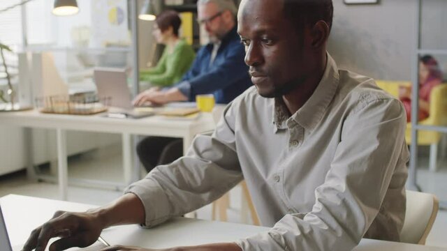 Afro-American man in casualwear using laptop and reading document while working at desk in office