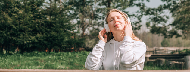 A girl in a white hoodie listens to music with headphones and relaxes sitting on a bench near a...
