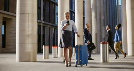 Young businesswoman with suitcase using smartphone waiting for taxi outside airport building