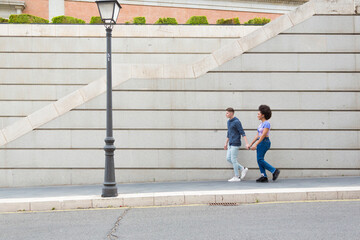 young multi-ethnic couple strolling hand in hand through city in blue casual clothes and a gray wall