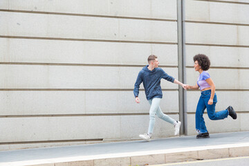 Fototapeta na wymiar young couple Caucasian man and African American woman running looking at each other in city with blue casual clothes and a gray wall