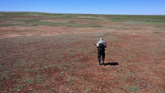 Tired soldier-sniper walks across the scorching desert land. View from the back.