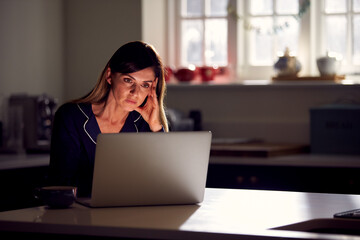 Unhappy Woman Wearing Pyjamas Sitting In Kitchen At Night Using Laptop