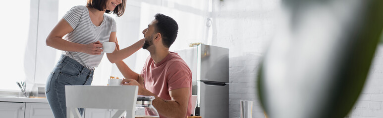 young couple gently touching each other and holding cups with coffee in kitchen, banner