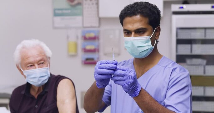 A Mid Shot Of An Indian Male Medical Nurse Administering A Covid-19, SARS-CoV-2 Vaccine Injection With A Needle and Syringe, To An Elderly Man. Wearing Safety Gloves And Protective Mask