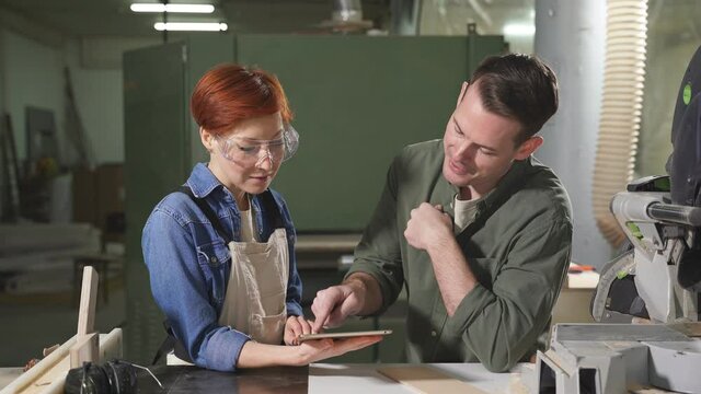 Young caucasian man and woman joiners using digital tablet while working in workshop, professional carpenters discussing working process, what to do, handmade and carpentry concept