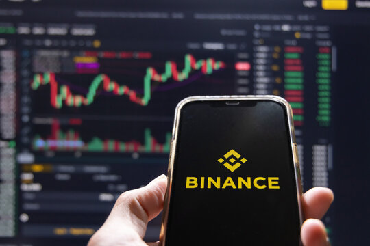 View of The Hand Holding a Mobilephone with Binance Logo on the Screen Against Bitcoin Graph Background