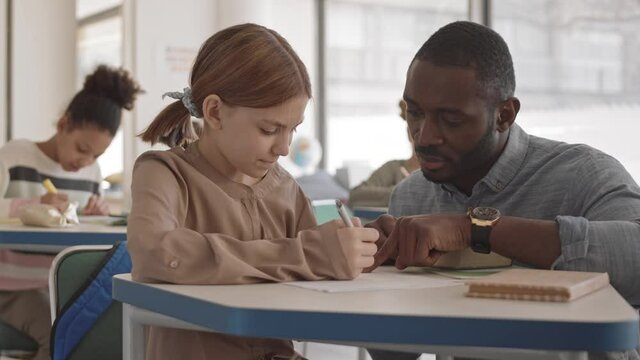 PAN medium shot of young male Afro American school teacher sitting on his laps next to smart 12-year-old girl doing test and explaining new material to her
