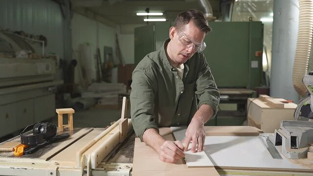 Male carpenter in green shirt wearing safety goggles for woodworking using pencil drawing line with measurement tool on wooden plank on table in workshop. Woodworking concept.