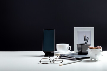 Office or home Office workspace. Desktop with smart phone template blank, notepads pens office suppliers cup of coffee drink plant flower. White work desk table on black background with copy space.
