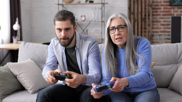 Two generation family. Senior mother and adult son playing video games using gamepads at home. Elderly versus youth. Confrontation of generations. Active modern elderly people.