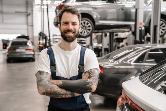 Bearded white car mechanic smiling while standing in garage indoors