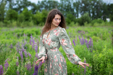 Girl walking on field on summer with wildflowers. Woman in long dress stands in blooming purple lupine field. Beautiful girl with wild flowers. Summertime. Summer vacation. Freedom and purity concept.