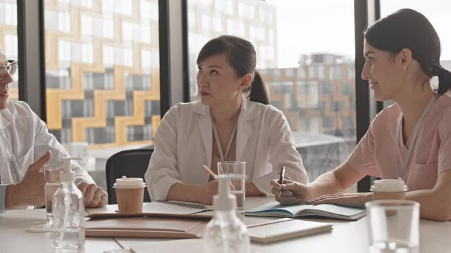 Medium PAN slowmo of contemporary male chief doctor talking to female colleagues sitting around table during medical conference or meeting