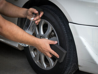 A man's hand holding a spray bottle of wheel cleaner and the other hand is holding a black sponge. wiping the edge of the tire