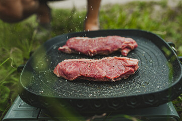 salt and pepper is sprinkled on meat steak is fried in grill pan on gas stove in open air on grass. cooking meal at picnic. healthy, full-fledged nutrition.delicious piece of meat. selective focus
