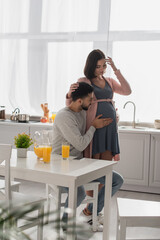 young man touching belly of smiling pregnant woman in kitchen