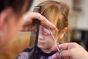 The hands of a hairdresser with a comb and scissors make a fashionable modern haircut for a girl for a child, a mother cuts a haircut for her daughter at home during the quarantine period
