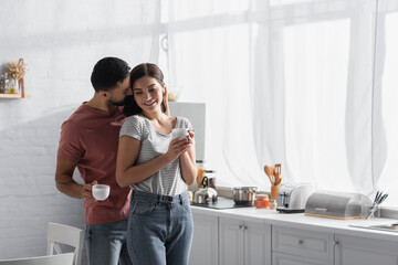 happy young couple hugging and kissing with coffee cups in kitchen