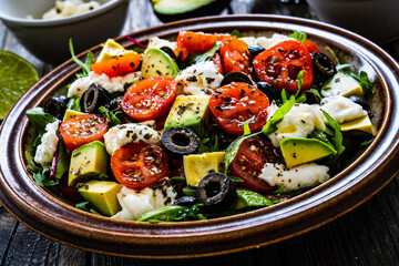 Fresh vegetable salad with mozzarella, tomatoes, avocado, lettuce, black olives and onion on wooden table
