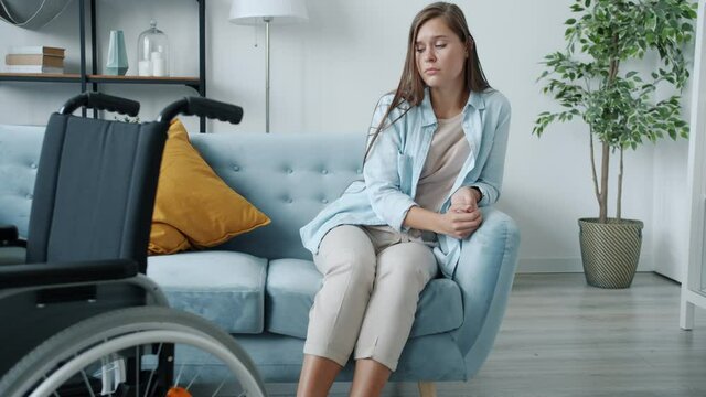 Portrait of unhappy disabled woman sitting on sofa alone and sighing with wheelchair visible in forefront. Depression and health problems concept.