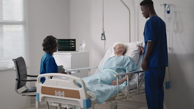 A team of african ethnic group doctors a man and a woman talk to an elderly male patient. A doctor and a nurse talk to a bedridden patient in a hospital ward