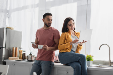 smiling young man standing with cellphone and coffee cup near girlfriend sitting with laptop at home