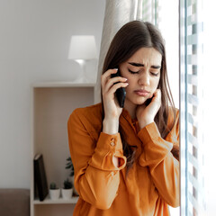 Cropped shot of a young business woman looking stressed using smart phone and standing near window. Sad woman receiving unwanted phone call. Alone thoughtful sadness woman using smartphone at home.