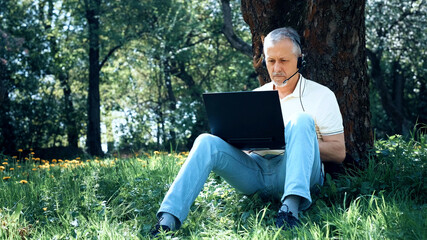 Obraz premium An elderly gray-haired man is working at a laptop, sitting on the grass among yellow flowers in a city park under a tree on a sunny summer day.