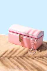 A pink pirates treasure chest box with some jewelry on sandy beach with palm leaf shadow, concept...
