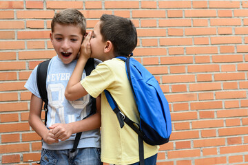 Two schoolboys, one is telling something in his ear and the other one opens his mouth in surprise