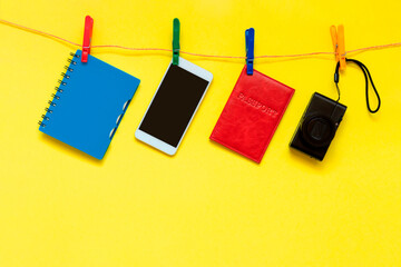 The concept of tourism is a set of things necessary for traveling: a camera, a passport, a phone and a notebook, suspended on clothespins on a yellow background.