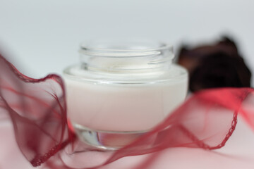 Closeup of a jar of moisturizer with red fabric and rose in the background.
