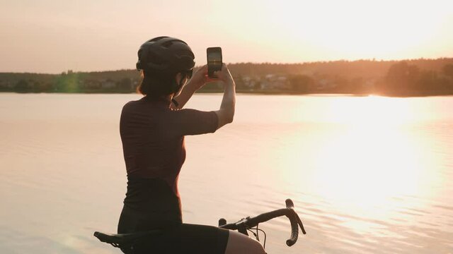 Woman cyclist standing on edge of coastline, looking at sunset and taking photos on smartphone